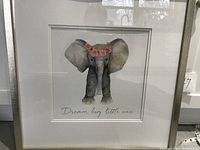 Frontal view of the framed watercolor print showing the full artwork and frame, confirming the subject as a baby elephant with a floral crown and text below.