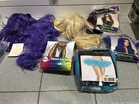 Photo showing multiple wigs in different colors and lengths including purple, blonde, and light blonde, some packaged wigs and a blue tutu accessory.