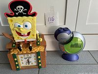Shows both clocks side by side on a surface. SpongeBob clock shaped like a treasure chest with character figure on top, night sky projector clock with buttons and LCD.