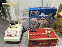 Osterizer blender with glass jar, Sunbeam Mixmaster Hand Mixer box, and Snoopy Sno-Cone Machine box together on a gray countertop.