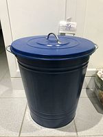 Large dark blue cylindrical metal storage bin with lid and side handles on tiled floor.
