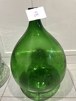 Front view of the large green glass demijohn carboy showing its bulbous shape and narrow neck with a tag number '2' attached.