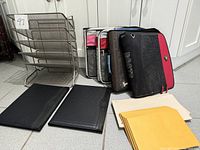 Photo showing two grey wire mesh shelves, four fabric binders of various colors including a 'ROOTS' branded binder, two black textured writing pads, manila file folders, and yellow envelopes arranged on the floor.