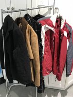 Five women's coats hanging on a rack showing variety of colors and styles including black, tan, plaid, and red gray winter jacket.
