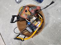 Top-down view of a round wooden bin filled with assorted power and hand tools including a cordless drill, reciprocating saw, tape measure, and other items.