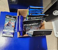 Sealed Panini basketball card packs arranged in a cardboard box on a blue surface, showing front of Contenders value packs and NBA Hoops 2020-21 packs