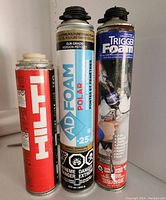 Three aerosol cans side by side showing front labels: HILT red can, AD FOAM POLAR white/blue, TRIGGER FOAM