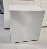 Closed white storage cabinet, cube shaped, showing the smooth front door surface without handles.