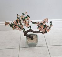 Photo showing a vintage Chinese bonsai tree with a twisted trunk, branches bearing glass and jade light peach blossoms and green leaves planted in a square ceramic pot with white pebbles.