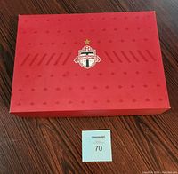 Closed Toronto FC branded red box with logo and star icon centered on lid, placed on hardwood floor with auction number card in foreground.