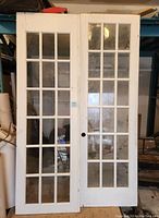 Two white painted solid wood French doors with multiple rectangular glass panel inserts arranged in a grid pattern, leaning upright against a wall indoors.