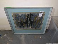 Full view of framed print leaning against wall on concrete floor, shows composition with three black horses and abstract background