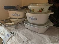 Five CorningWare dishes stacked, including two rectangular and one square dish with blue cornflower pattern, one small bowl with vegetable pattern, and stacked lids and metal holders beside them.