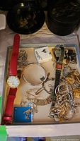 Overview of assorted costume jewelry pieces spread out in a box including two watches, bracelets, brooches, cufflinks.