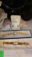 Evergold 10K earrings in display box with two ladies watches in separate boxes on a white surface. Watches have rectangular faces with yellow stone bracelets, one amber, one tiger eye.
