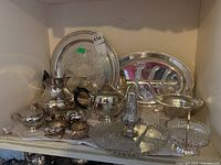 Overview of silver plated tea set items arranged on shelf, including serving trays, teapots, bowls, and glassware.