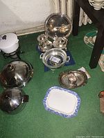 Photo shows assortment of silver plate items including trays, teapots or creamers, and a covered bowl on green carpet with some ceramic pieces nearby.