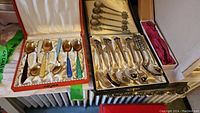 Overview of two open boxes containing multicolor enamel spoons on left and decorative silver-tone spoons on right.