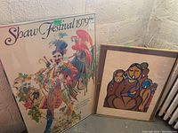 Wide view of both framed prints leaning against a wall showing the Shaw Festival 1979 poster with broken glass and second colorful native art print