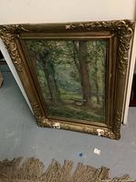 Full view of vintage framed oil painting showing a wooded landscape with a stone bench, frame visible with damage.