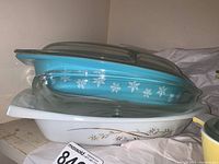 Two Pyrex dishes stacked; lower dish white with gold pattern, upper dish blue with white snowflakes and clear glass lid on top.