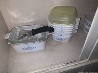 Shelf with white casserole dish with blue floral pattern, clear glass lid with black handle, and a stack of plastic lids