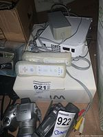 Nintendo Wii console with connected power adapter and AV cables on top of original Wii box, two Wii Remote controllers placed beside console.