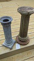 Photo shows two garden pedestals placed on a wooden deck. The pedestal on the left is shorter with a square base and hollow circular top. The pedestal on the right is taller, with a round base and a wider round top surface, both pedestals have fluted vertical columns and visible weathering.