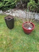 Outdoor scene showing both planters placed on grass, one tall brownish floral-shaped planter and one red round planter with plant remnants inside.