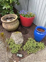 Three outdoor planters together showing red planter with purple flowers, blue planter with dirt, natural clay planter with dried plants.