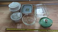 Seven bakeware items arranged on floor including Pyrex and CorningWare casseroles, glass baking dishes, clear bowl with lid, and green ironware casserole with lid.