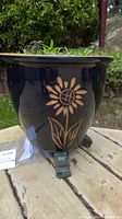 Side view of black ceramic planter showing raised sunflower design and small decorative feet.