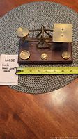 Small brass and wood balance scale placed on a wooden table next to a measuring tape showing about 7 inches width. Wooden base supports brass balance beam with a dial and round weighing pan. Three cylindrical brass weights are positioned on the base.