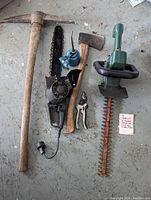 Photo showing pickaxe with wooden handle, Mastercraft 14 inch chainsaw with cord, small blue oil can, wooden handled axe, small hand clippers, and Black & Decker green and black 18 inch electric hedge trimmer with dual-sided blade.