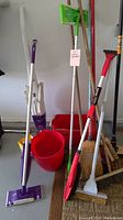 Multiple cleaning tools including Bissell Easy Vac, brooms, mop buckets, dusters, and dustpans arranged against a wall