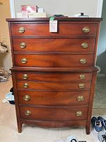 Front view of Dixie highboy dresser showing stacked drawers, brass oval handles, scalloped base, and smooth polished brown wood finish.