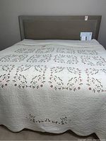 Full view of queen quilt on a bed showing white quilt with embroidered pink and green floral patterns.