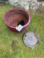 Top-down view of large red ceramic planter with soil inside and saucer beside it on grass