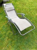 Photo of the entire zero gravity lounge chair on grass showing metal frame, beige fabric seat, padded headrest and plastic armrests.