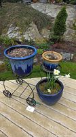 Shows three blue ceramic planters arranged outdoors on a table, two on black wire stands, one on table surface containing growing plants