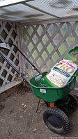Green Scotts Turf Builder Mini spreader with large black wheels and black handle holding a bag of Scotts Turf Builder Pro Lawn Food fertilizer inside, placed in a greenhouse corner.