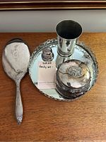 Photo showing the full silver vanity set: hand mirror, tray, powder box, cup, and shaker on wooden top.