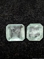 Close-up of two square Colombian emerald gemstones showing facets and light green color with inclusions on a dark fabric background.