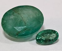 Two green oval emerald stones on white background, one larger rough/natural and one smaller faceted.