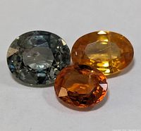 Three oval faceted sapphires in blue-gray, yellow, and orange colors on white background.