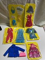 Lot of Barbie doll clothes including various dresses, pants, coats, handbags, all on yellow backing cards in original plastic packaging.