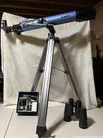 Full view of blue Sky-Watcher telescope mounted on aluminum tripod, with accessories box and binoculars placed on floor