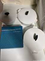 Three white Corning Ware buffet servers with lids and black knobs arranged on table next to candle warmer box.