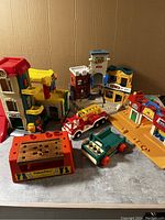 Group photo of vintage Fisher Price toys including playsets and vehicles on gray surface against brown background.