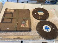 Three items laid flat: two metal 16mm film reels with visible labels and one large worn cardboard 16mm film carrier secured by olive green canvas straps and metal buckles.